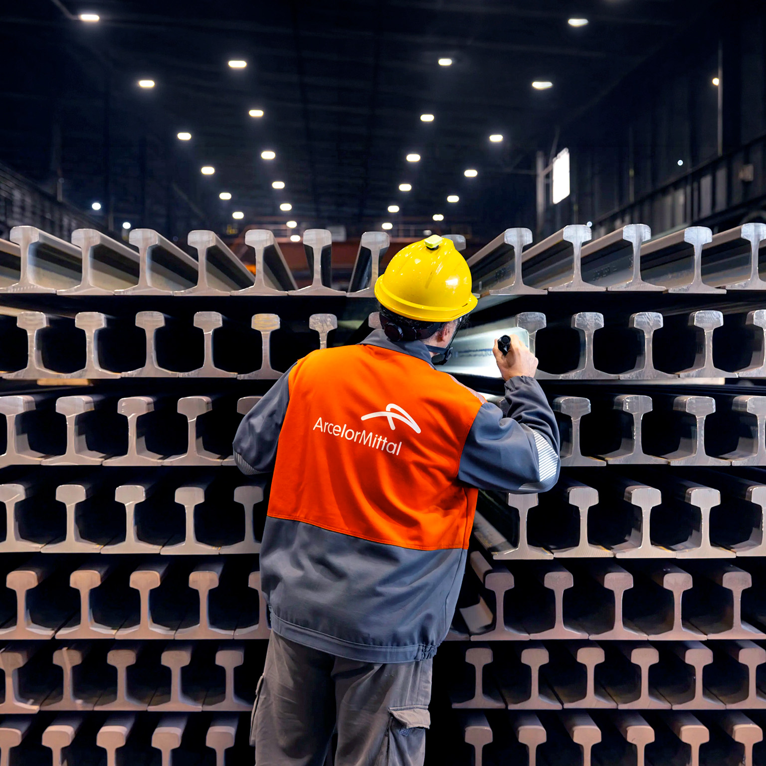 ArcelorMittal employee inspecting stacked steel railway rails inside a manufacturing facility.