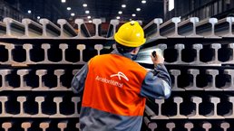 ArcelorMittal employee inspecting stacked steel railway rails inside a manufacturing facility.