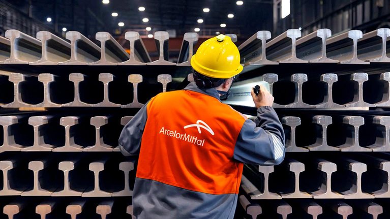 ArcelorMittal employee inspecting stacked steel railway rails inside a manufacturing facility.