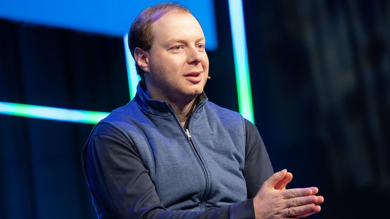 Andrey Khusid speaking on stage with a headset microphone, gesturing with his hands against a dark background accented by blue and green light strips.