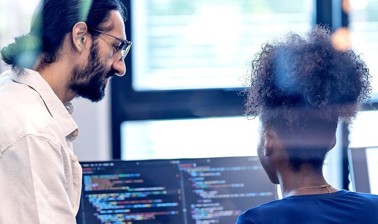 Two software developers collaborate at a workstation, reviewing code displayed on dual monitors in a bright office setting.