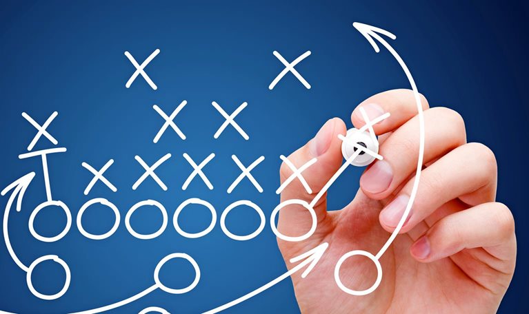 A football play diagram is being sketched by a hand, featuring Xs and Os to represent players and arrows indicating their movements.