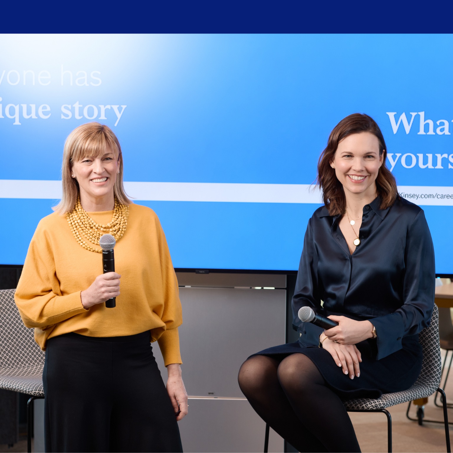 Marie and Blair at some event; Marie sitting at a stool, Blair standing and holding a microphone; both smiling.