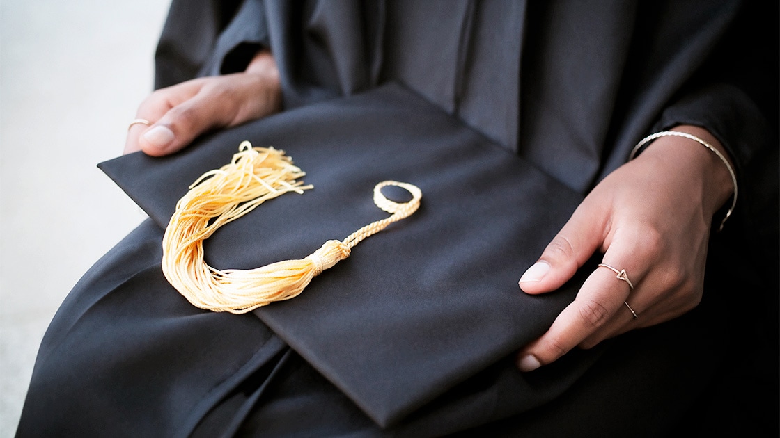 A close-up of the hands of a Black college student who is wearing a graduation gown and holding a graduate cap with a yellow tassel.