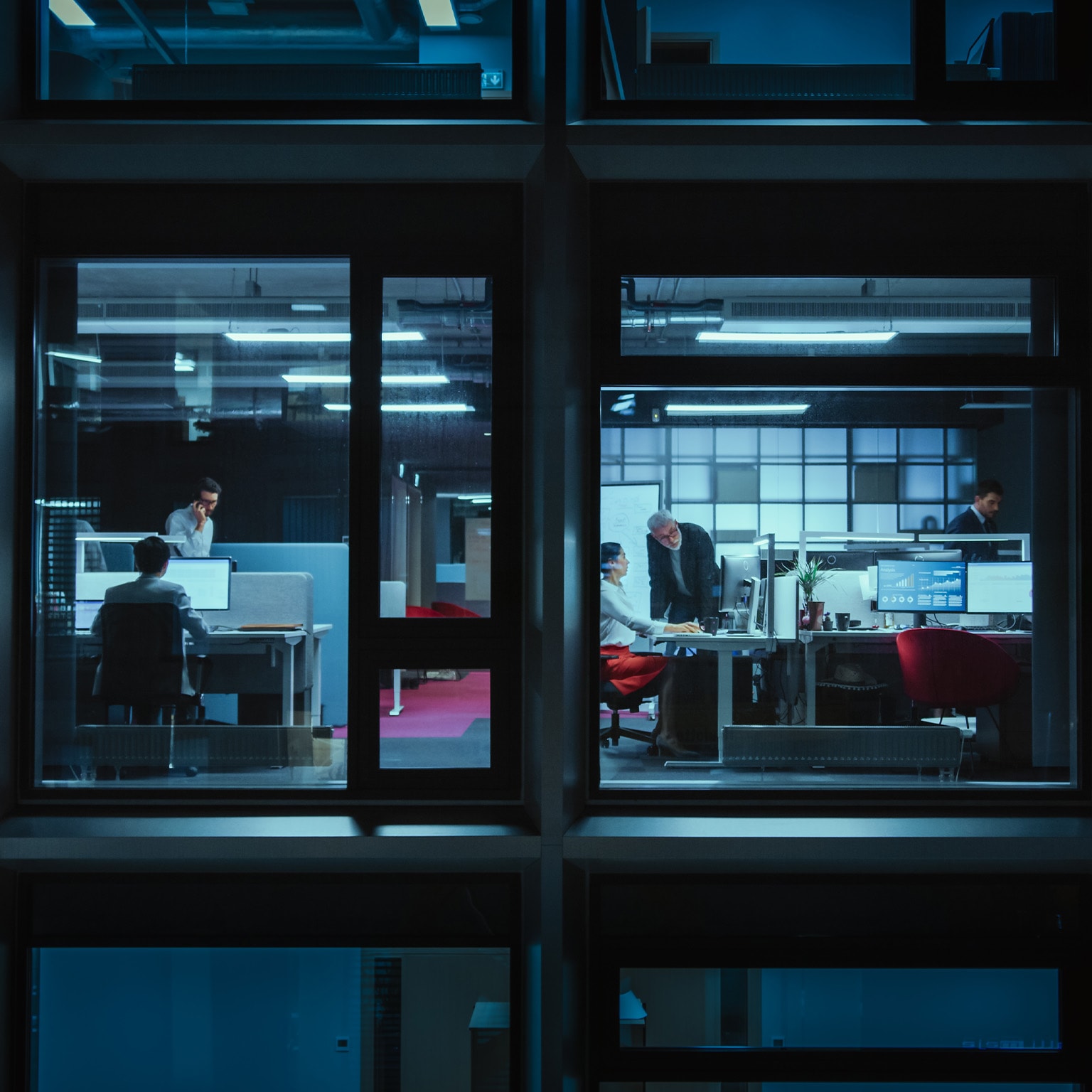 Shot from outside the window businessmen and businesswomen in the office doing financial business in the evening working on computers and delegate tasks. - stock photo