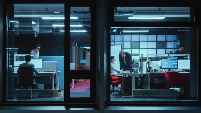 Shot from outside the window businessmen and businesswomen in the office doing financial business in the evening working on computers and delegate tasks. - stock photo