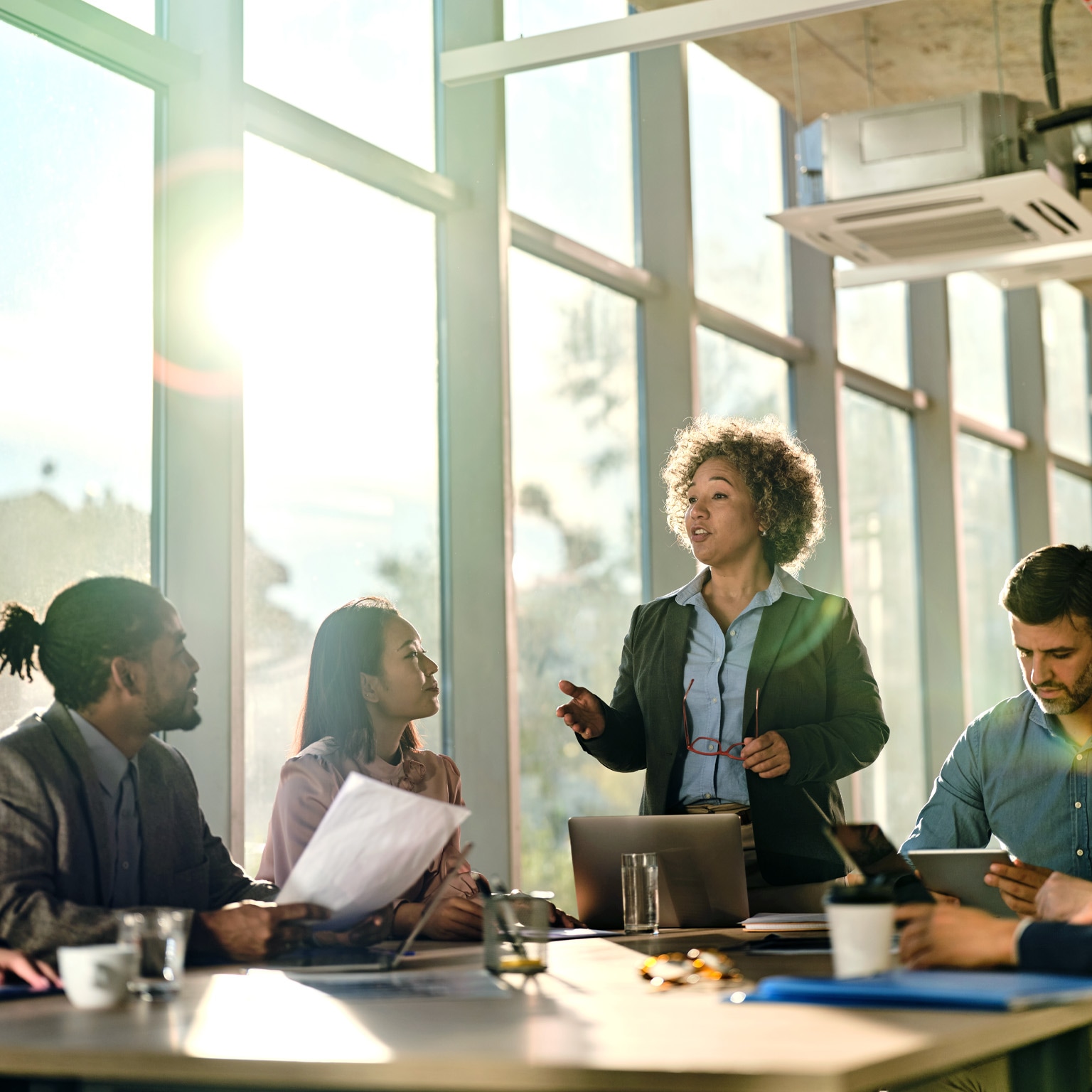  At the front of the conference room table, a woman is leading a meeting while a wall of large windows behind her allows light to stream in and cast long shadows over the table.