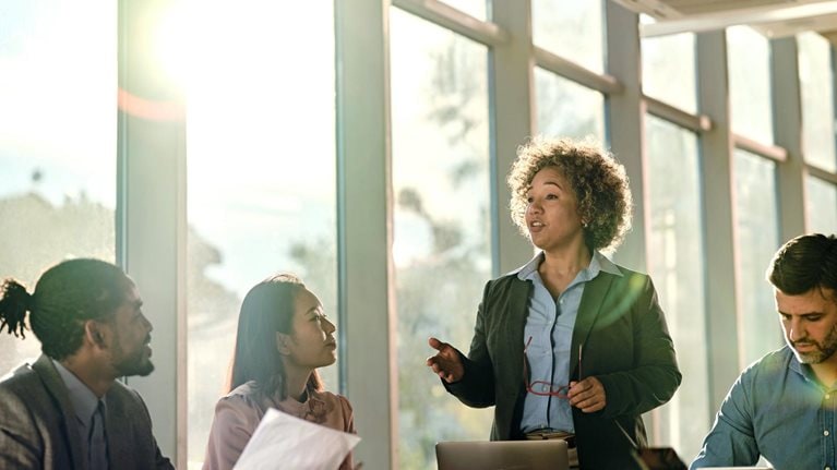 At the front of the conference room table, a woman is leading a meeting while a wall of large windows behind her allows light to stream in and cast long shadows over the table.