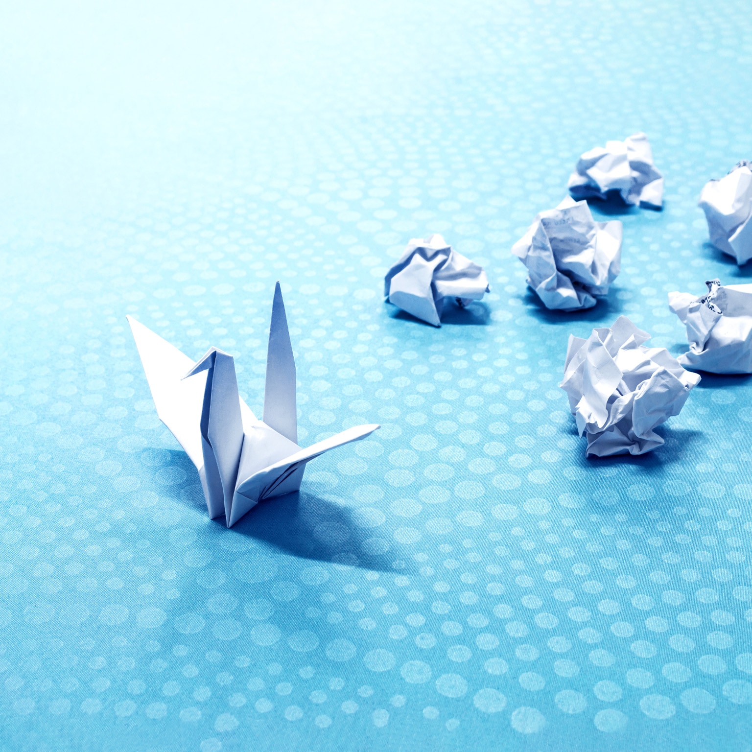 A lone origami crane sits on a blue background with white polka dots. Behind the crane is a cluster of crumpled balls of white paper.