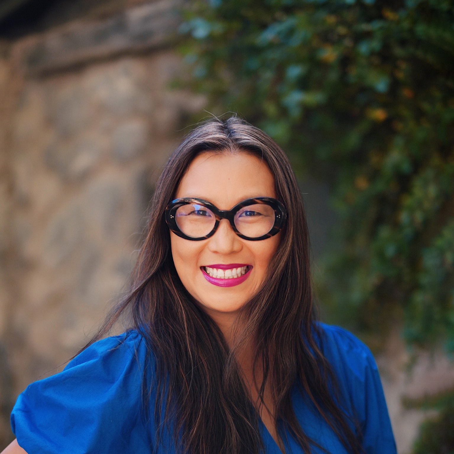 Portrait of a smiling Bonnie Wan with a stone wall and greenery blurred in the background. 