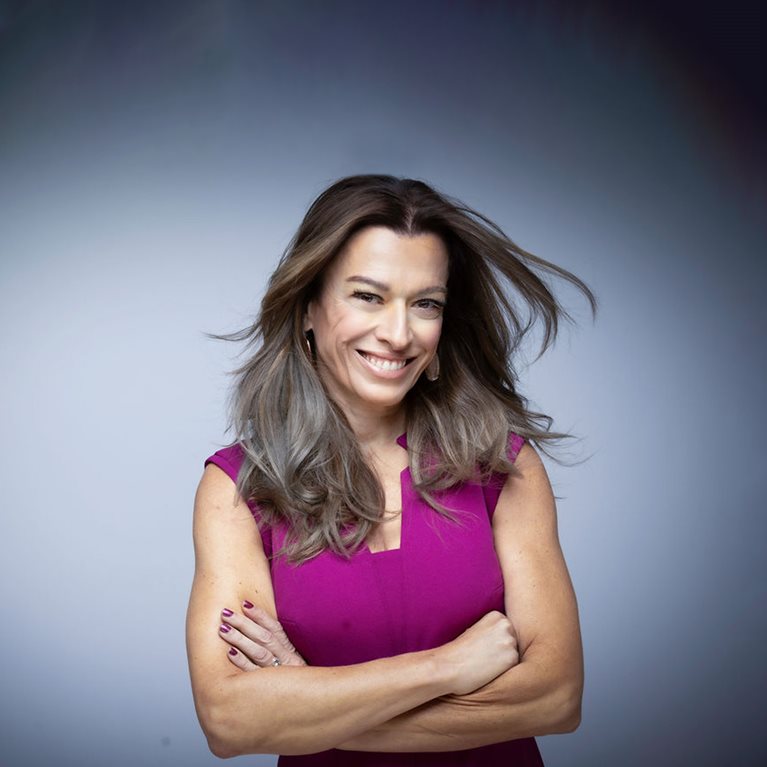 A portrait of Anne Marie Chaker smiling, with flowing brown hair, wearing a sleeveless purple dress. The background is a plain light gray or blue.