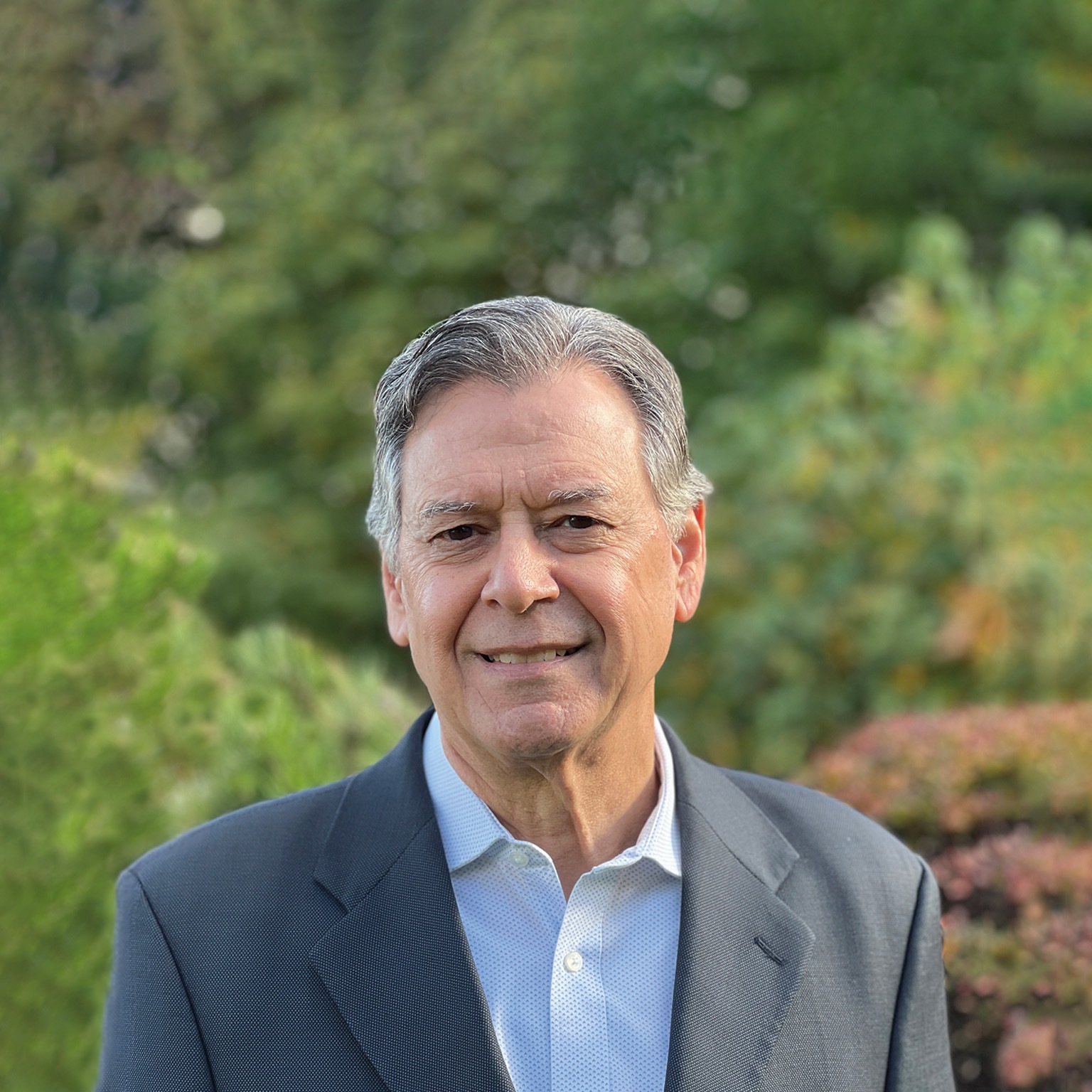 Lawrence Ingrassia with a slight smile and wearing a gray suit jacket and collared shirt, with lush outdoor scenery in the background.