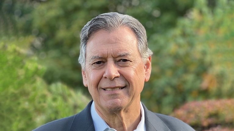 Lawrence Ingrassia with a slight smile and wearing a gray suit jacket and collared shirt, with lush outdoor scenery in the background.