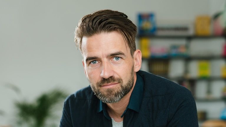 Mark Tuitert is depicted in a portrait wearing a navy blue overshirt with a collar, seated and leaning forward with evident interest. His hair is styled in a sleek side sweep and he sports a well-groomed beard.