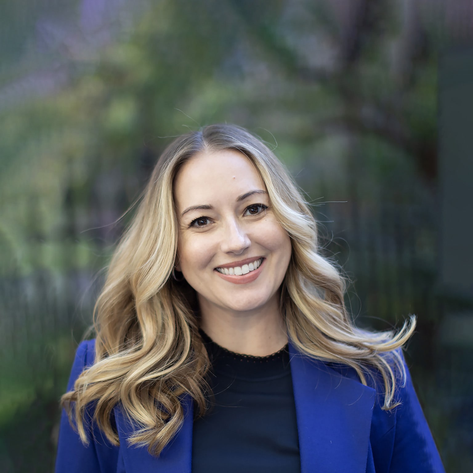 A portrait of Tessa West, wearing a royal blue blazer and smiling, with a blurred background of greenery.