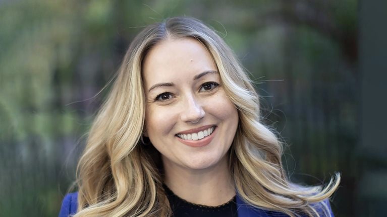A portrait of Tessa West, wearing a royal blue blazer and smiling, with a blurred background of greenery.
