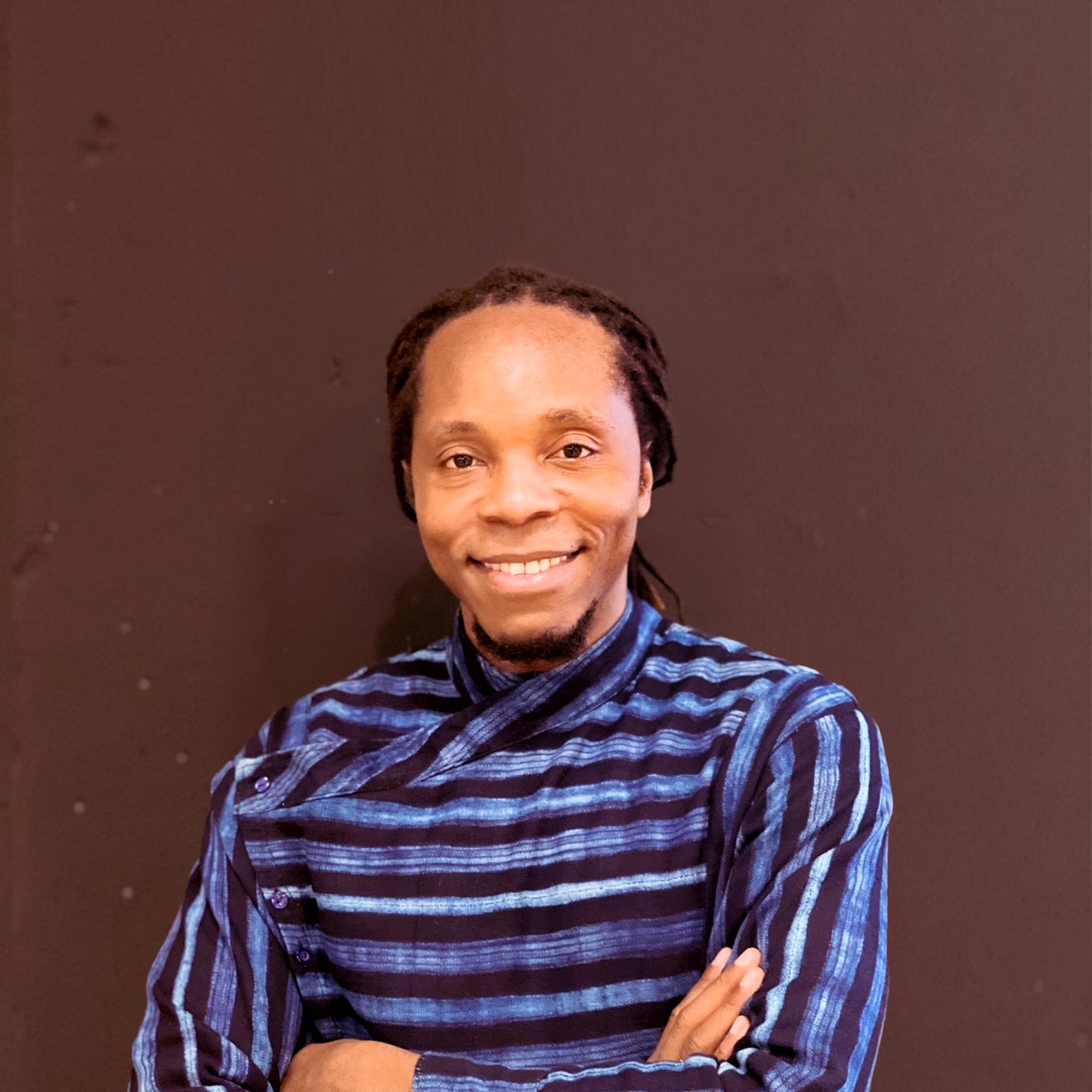 Portrait of author David Moinina Sengeh. Sengeh has a friendly smile and is wearing an artistic long sleeve blue and black striped shirt and photographed against a brown wall.