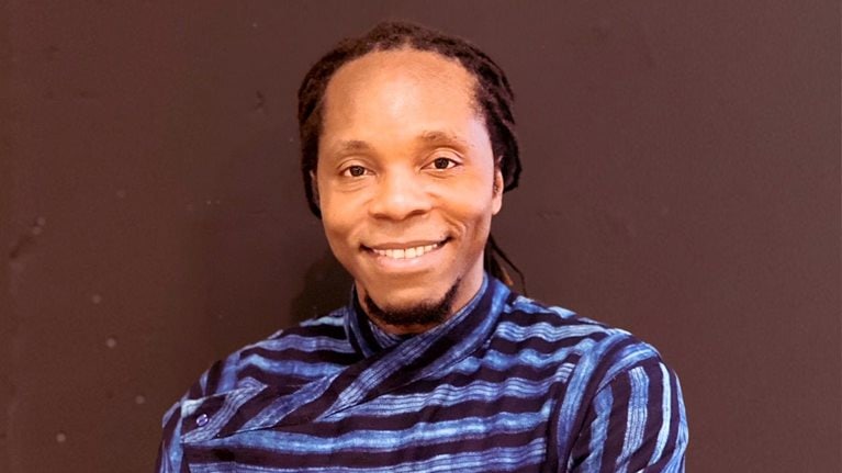 Portrait of author David Moinina Sengeh. Sengeh has a friendly smile and is wearing an artistic long sleeve blue and black striped shirt and photographed against a brown wall.