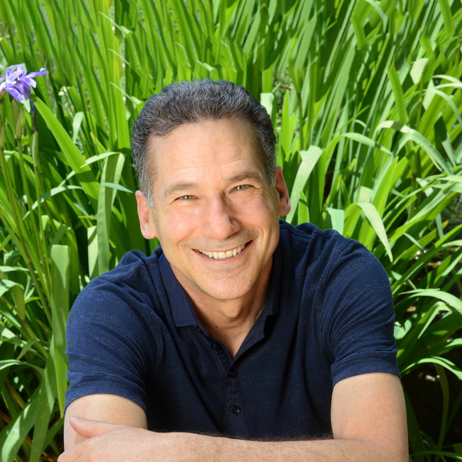 A portrait of William Ury in a navy blue collared shirt against a backdrop of lush greenery.