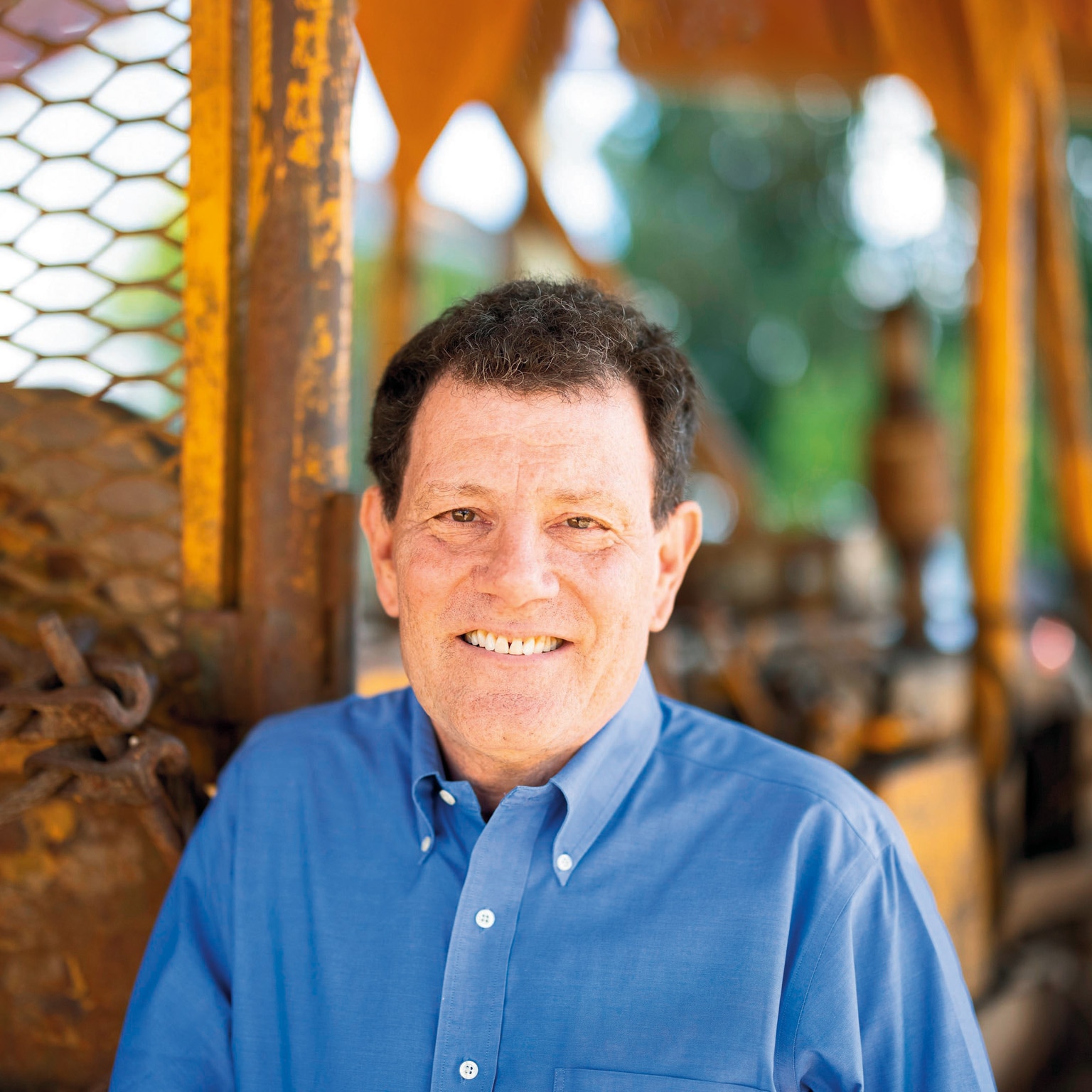 A photograph of a smiling Nicholas Kristof wearing a blue button-down shirt against a backdrop of weathered metal machine.