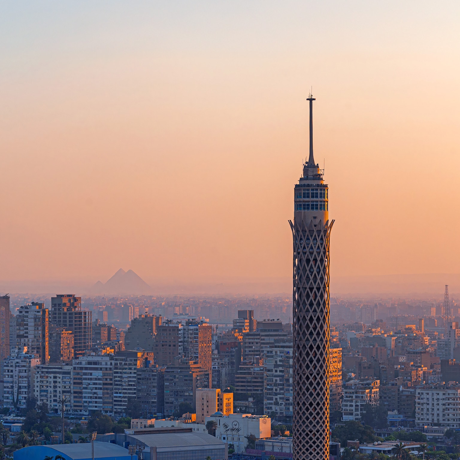 In the golden glow of sunset, Cairo Tower rises above Gezira Island while, in the hazy distance, a Great Pyramid of Giza faintly appears—bridging ancient history and modern Cairo in one quintessential view.