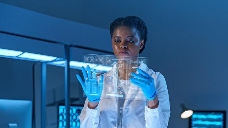A serious female scientist in a lab coat and protective gloves stands in a modern lab, working on a small HUD or graphic display