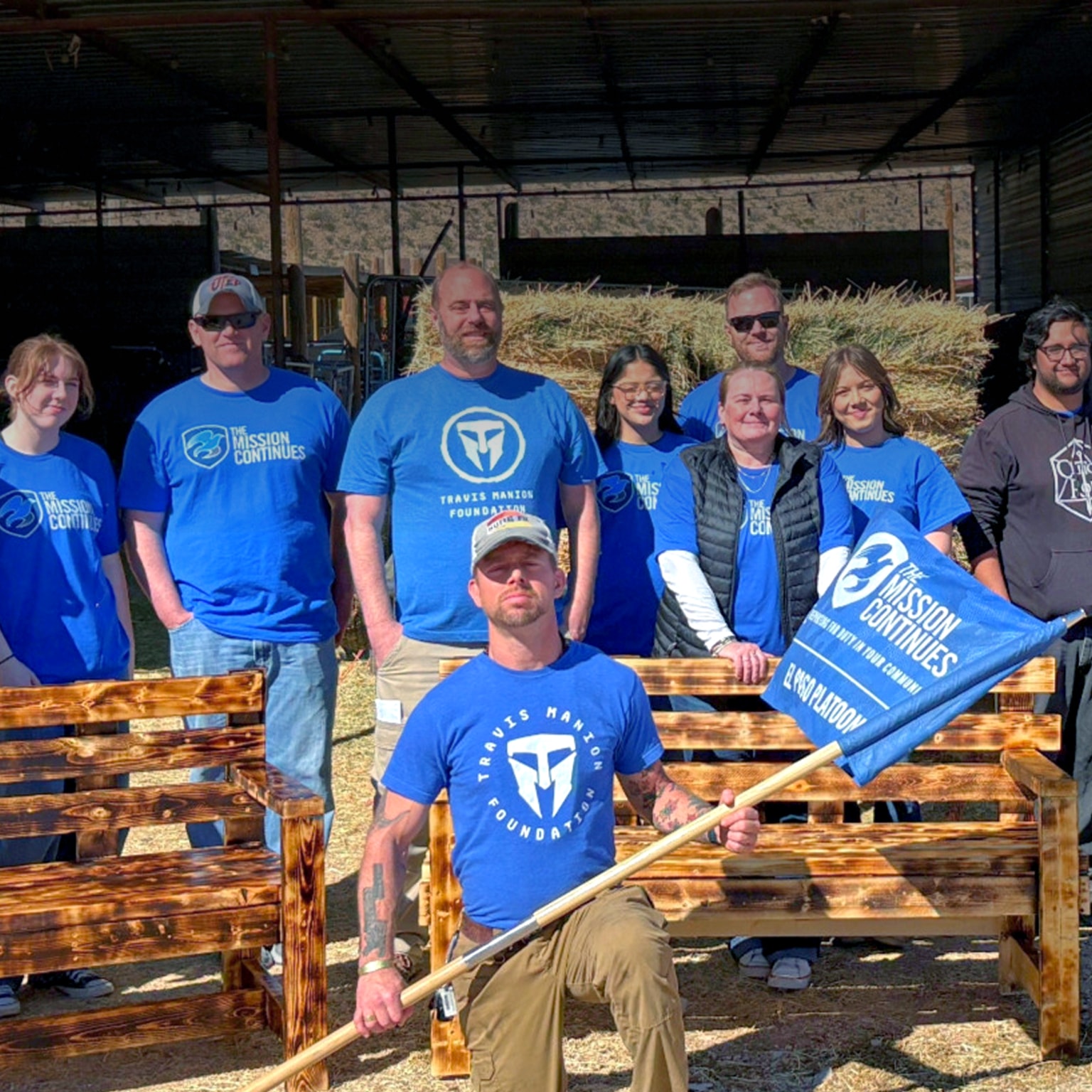 The Mission Continues’s volunteers posing outdoors behind newly built wooden benches, with one person kneeling in front holding a flag.