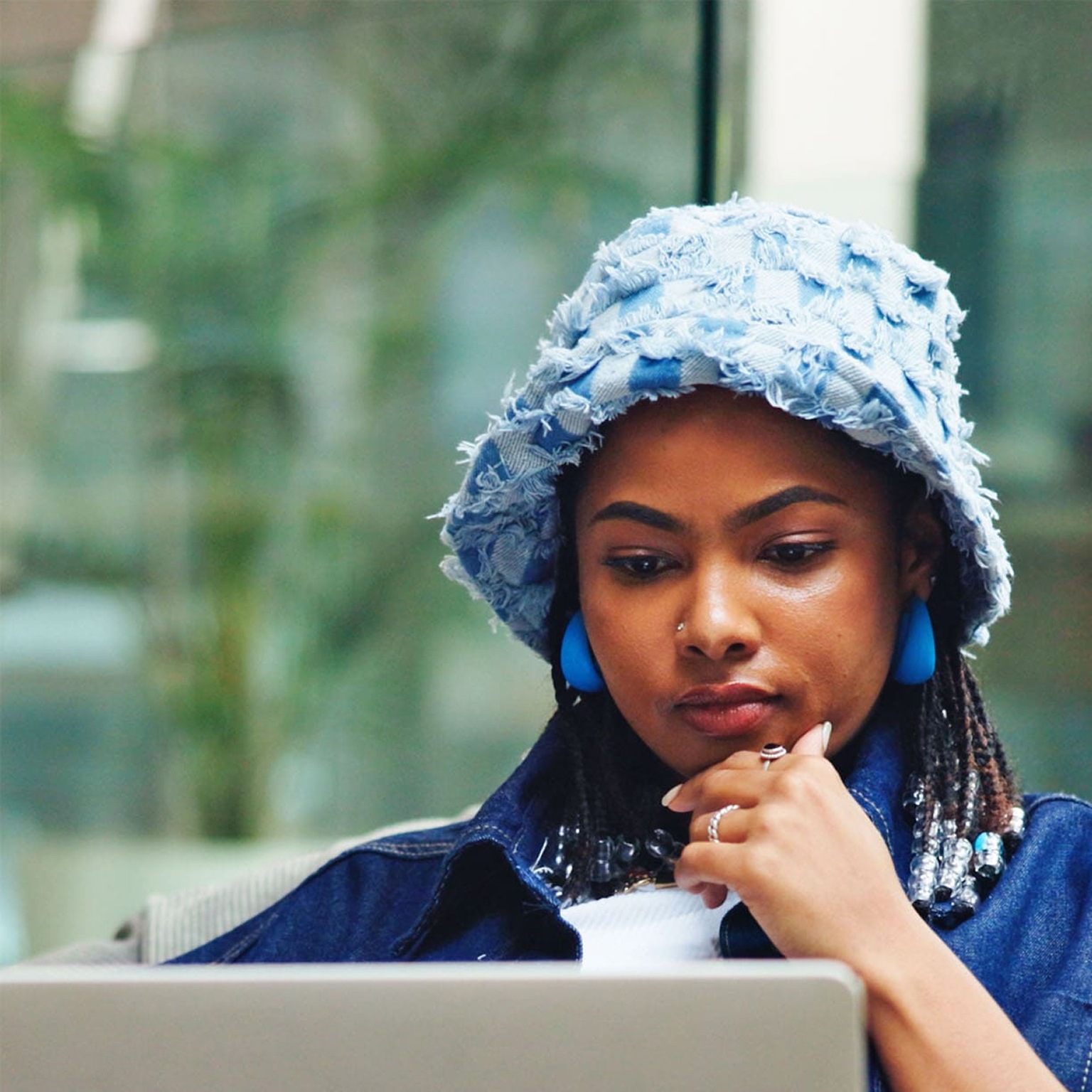 A person wearing a light blue textured bucket hat and blue earrings sits on a cushioned bean bag, concentrating on a laptop balanced on their lap with one hand resting near their chin. The background shows a modern, bright indoor space with glass walls and blurred furnishings.