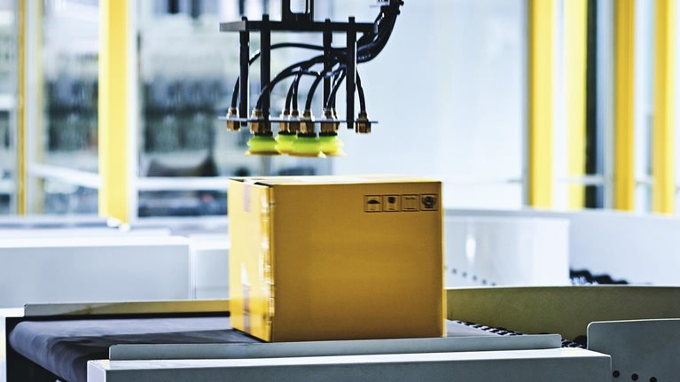 Close-up of robotic arm carrying cardboard box on conveyor belt at modern distribution warehouse.