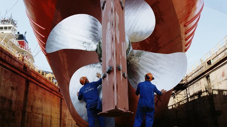 The image captures workers performing maintenance on a large ship's propeller and rudder in a dry dock. This process involves essential inspections and upkeep.