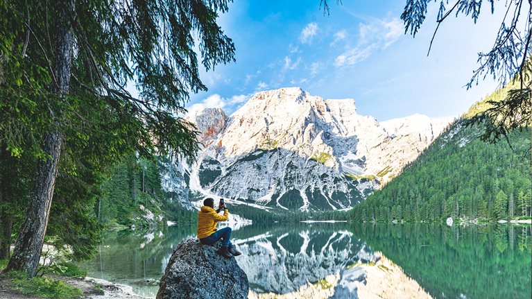 Hiker taking picture of mountains, enjoying sunrise sitting on rocks on shore of Lake Braies. - stock photo