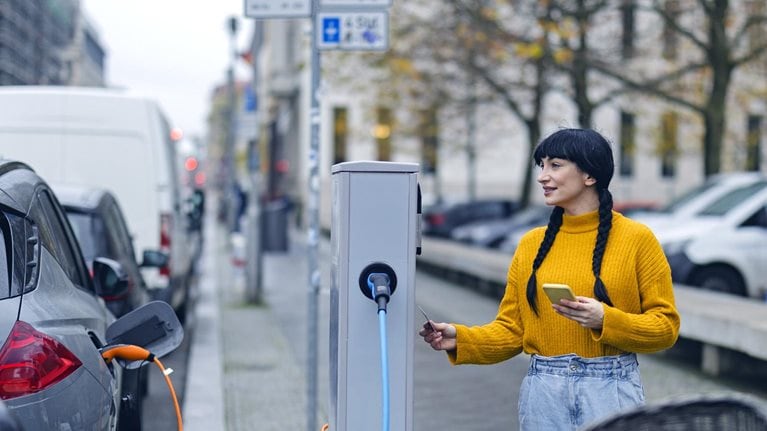A woman with smartphone and credit card preparing to pay at an electric vehicle charging station on a tree-lined European city street.
