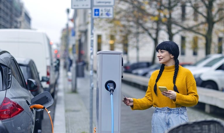 A woman with smartphone and credit card preparing to pay at an electric vehicle charging station on a tree-lined European city street.