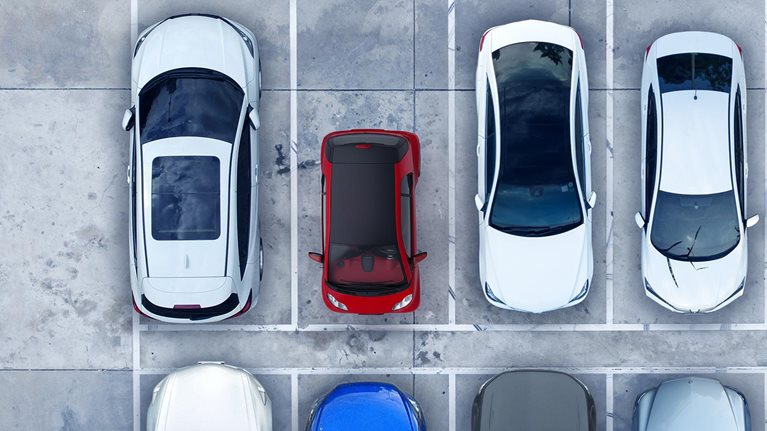 An overhead view of a red, ultra-light microcar parked among 7 full size vehicles highlighting the contrasts in size and space between them