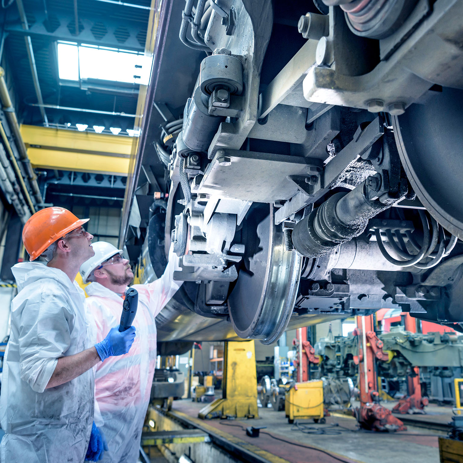 Two workers wearing hard hats and protective clothing inspect the wheel assembly and undercarriage of a train inside an industrial maintenance facility, with one worker pointing at a component while the other holds an inspection device.