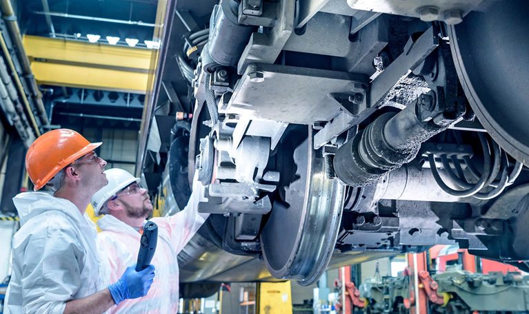 Two workers wearing hard hats and protective clothing inspect the wheel assembly and undercarriage of a train inside an industrial maintenance facility, with one worker pointing at a component while the other holds an inspection device.