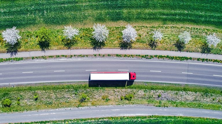 Aerial view of a red and white semi-truck driving down an interstate road though green farm fields