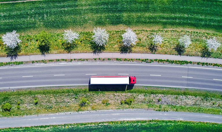 Aerial view of a red and white semi-truck driving down an interstate road though green farm fields