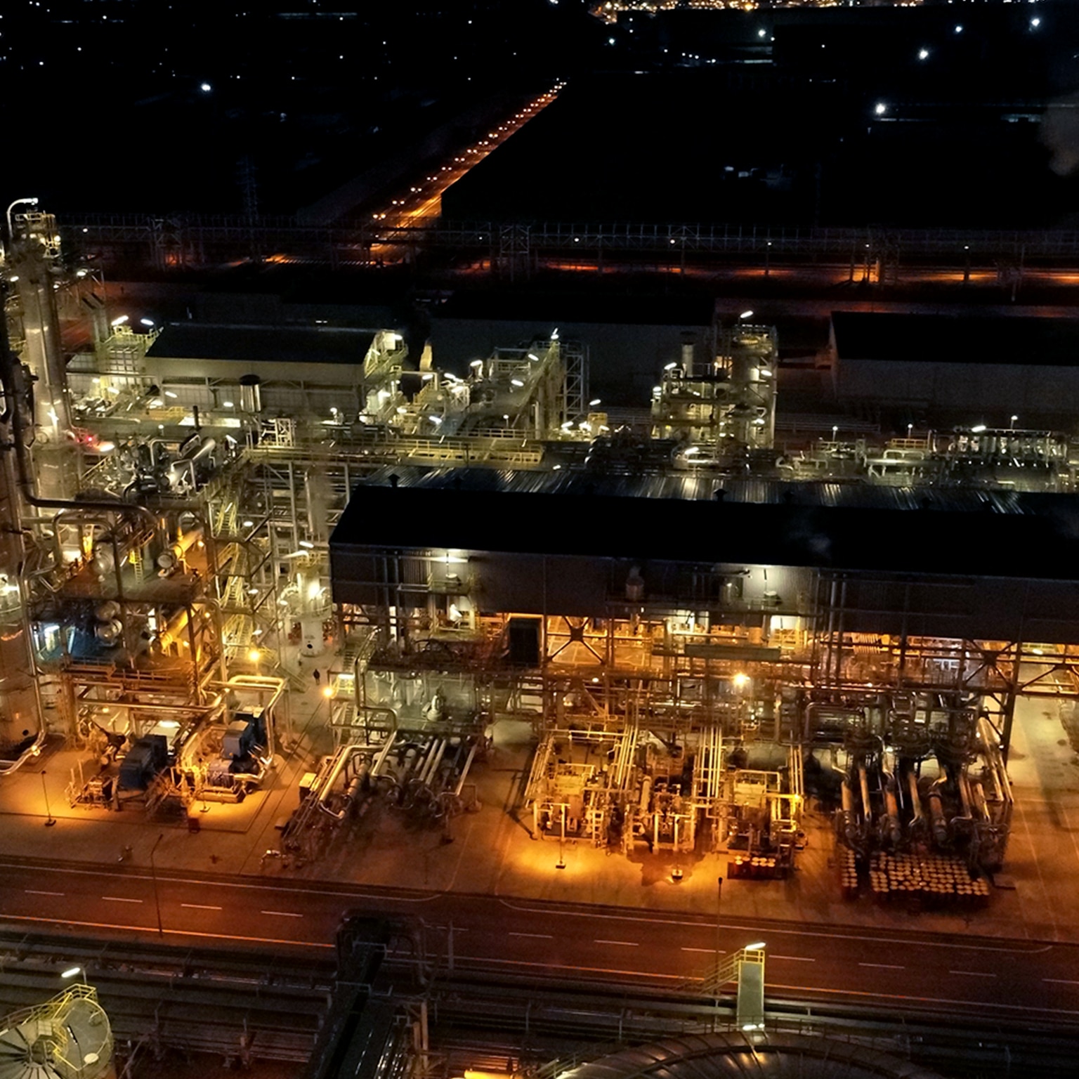 Aerial nighttime view of an illuminated industrial processing facility with interconnected buildings, pipelines, and storage tanks.