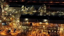 Aerial nighttime view of an illuminated industrial processing facility with interconnected buildings, pipelines, and storage tanks.