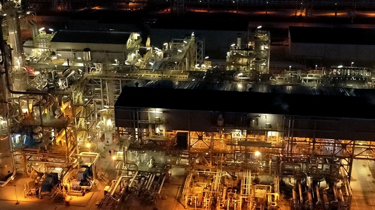 Aerial nighttime view of an illuminated industrial processing facility with interconnected buildings, pipelines, and storage tanks.
