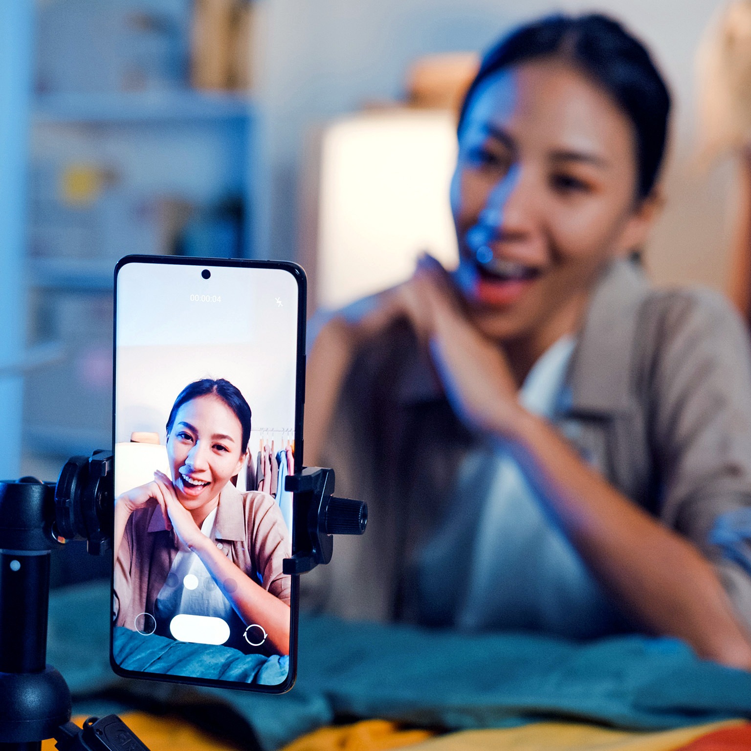 Asian woman live-streamed ecommerce sell clothes in home at night. Beautiful girl using the smartphone and tablet for recording video.