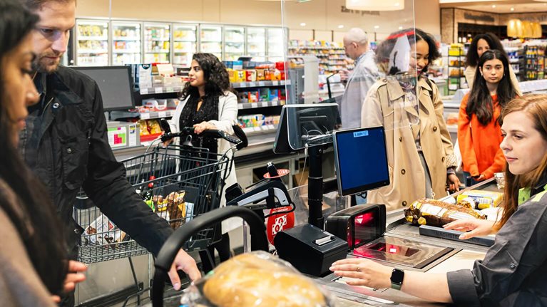 This image depicts a busy scene at a supermarket checkout counter. A cashier is scanning groceries for a customer, with other shoppers waiting in line behind them.