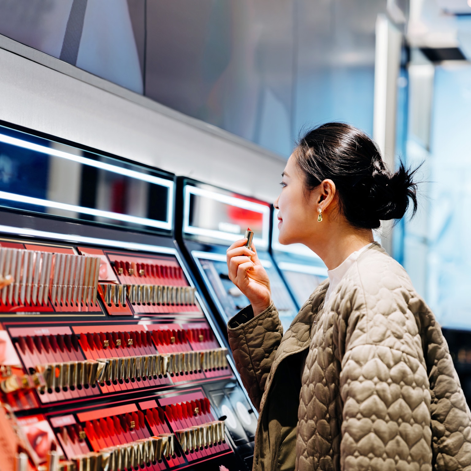 A woman applying lipstick at a makeup counter. The counter has rows of lipsticks in various shades of red and pink, and is lit by bright lights.