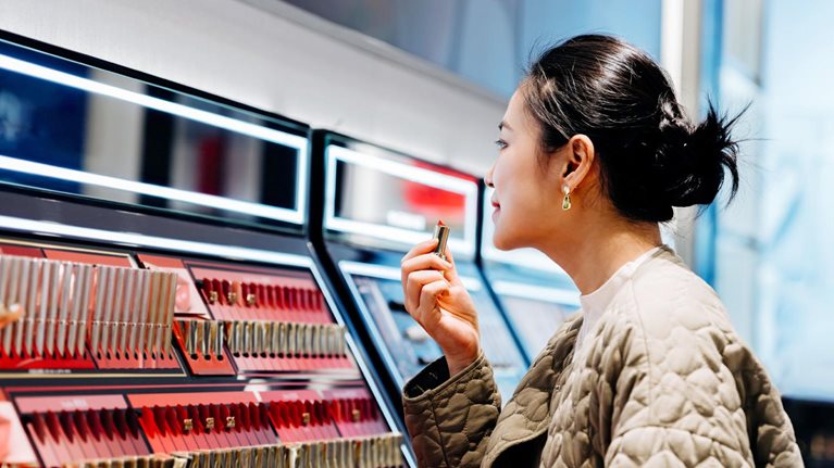 A woman applying lipstick at a makeup counter. The counter has rows of lipsticks in various shades of red and pink, and is lit by bright lights.