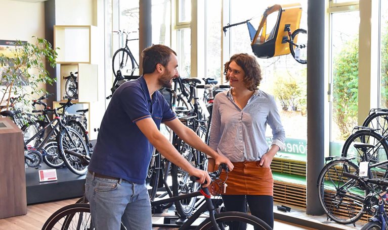 A man leaning on the handlebars of a bike in a brightly lit bike shop while a smiling store employee looks on. 