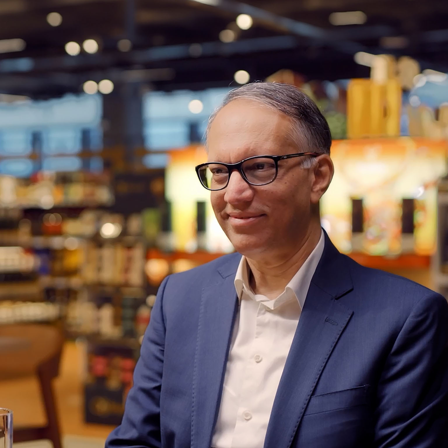 Man in a blue suit and glasses sits in a warmly lit store or café setting, smiling slightly, with shelves and lights blurred behind.