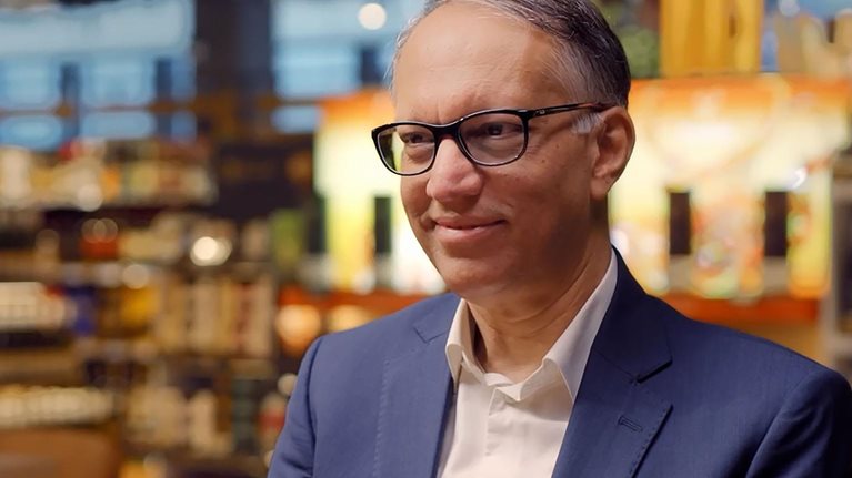 Man in a blue suit and glasses sits in a warmly lit store or café setting, smiling slightly, with shelves and lights blurred behind.
