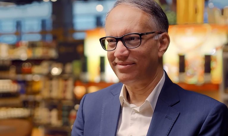 Man in a blue suit and glasses sits in a warmly lit store or café setting, smiling slightly, with shelves and lights blurred behind.
