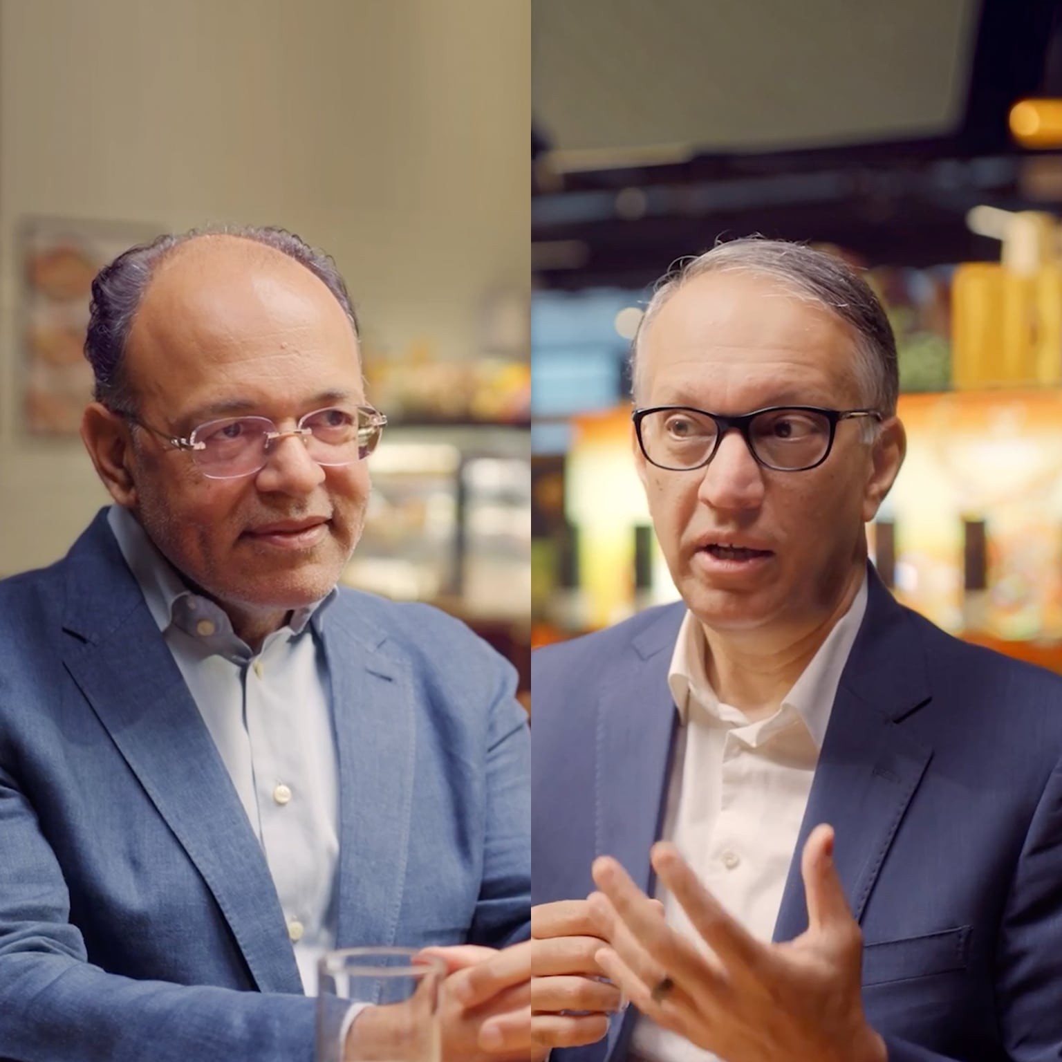 Vipul Chawla in a blue suit and glasses sits in a warmly lit store or café setting, smiling slightly, with shelves and lights blurred behind.